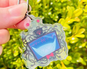 May include: A clear acrylic keychain featuring a cartoon character with a black and blue television screen face, red bow tie, and black wires. The character has a large, toothy grin and a heart rate monitor on the screen.