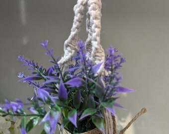 May include: A white macrame plant hanger with a burlap pot filled with purple flowers.