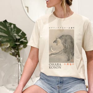 May include: A woman wearing a cream colored t-shirt with a black and white illustration of a tiger and a crescent moon. The text "OHARA KOSON" and Japanese characters are printed on the shirt.