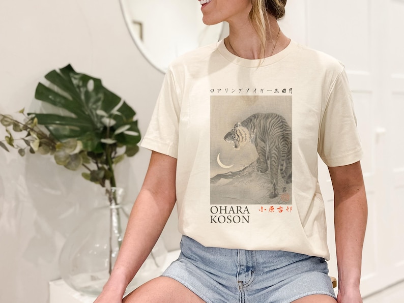 May include: A woman wearing a cream colored t-shirt with a black and white illustration of a tiger and a crescent moon. The text "OHARA KOSON" and Japanese characters are printed on the shirt.