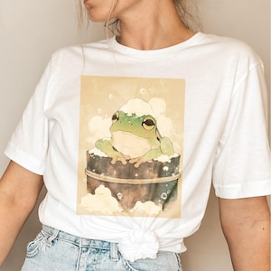 May include: White t-shirt with a graphic of a green frog in a wooden tub with bubbles. The frog is wearing a white foam hat.