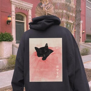 May include: Black hoodie with a graphic print of a black cat sleeping on a pink blanket. The Japanese text on the print says "おやすみ" which means "good night".