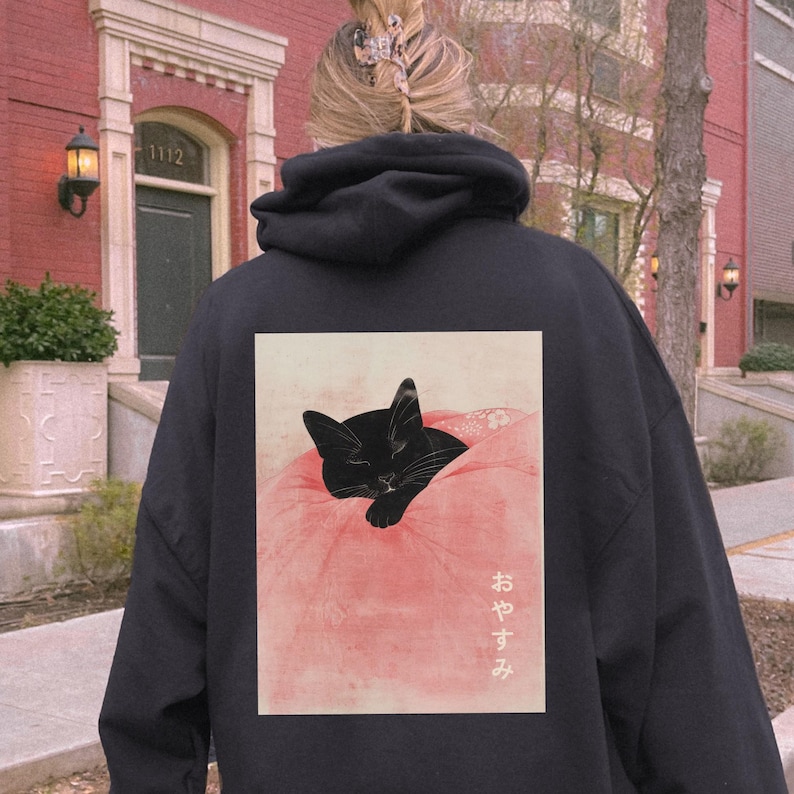 May include: Black hoodie with a graphic print of a black cat sleeping on a pink blanket. The Japanese text on the print says "おやすみ" which means "good night".