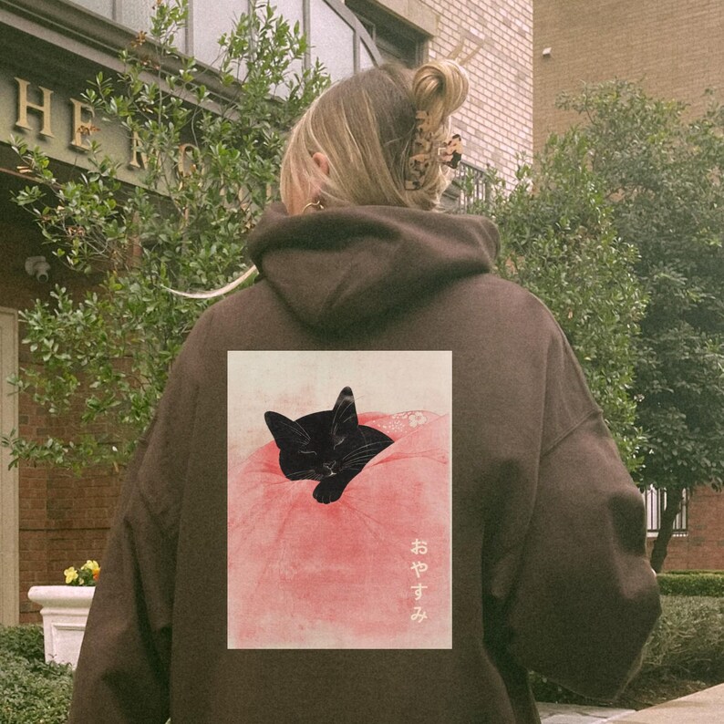 May include: A brown hoodie with a black cat sleeping on a pink pillow printed on the back. The Japanese text "おやすみ" is printed below the cat.