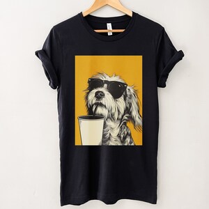 May include: Black t-shirt with a graphic of a dog wearing sunglasses and drinking from a cup with a straw. The dog is on a yellow background.