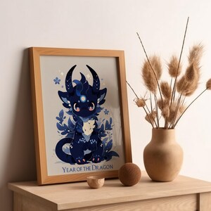 Pu&ograve; includere: Una stampa incorniciata con un drago di cartone animato blu e nero con una luna sulla fronte. Il drago &egrave; circondato da fiori e foglie blu. Il testo "Anno del Drago" &egrave; in fondo alla stampa.