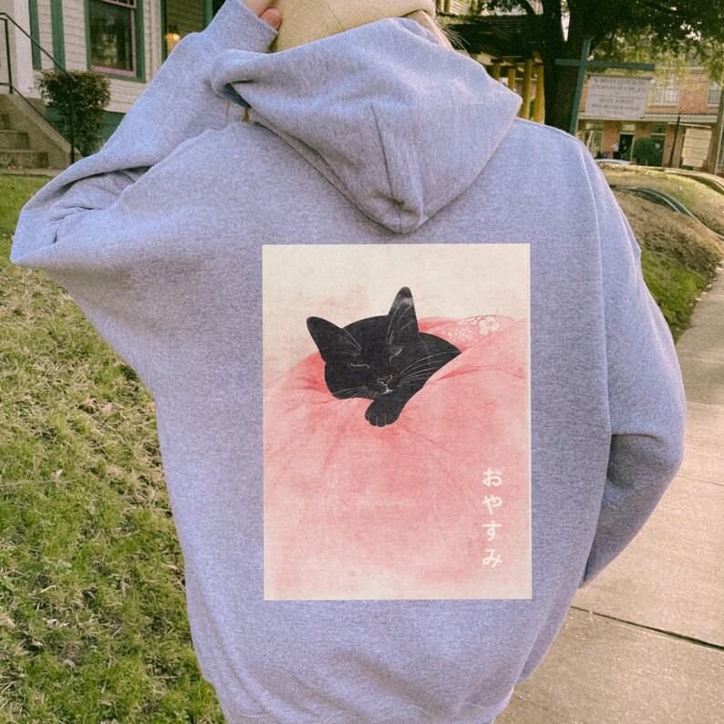 May include: A gray hoodie with a black cat sleeping on a pink pillow. The cat is in a Japanese-style illustration. The text "おやすみ" is printed on the hoodie.