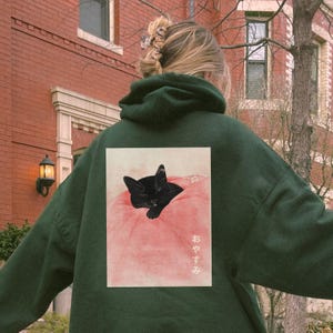 May include: A dark green hoodie with a black cat sleeping on a pink pillow. The cat is surrounded by flowers and the text "おやすみ" is printed on the pillow.