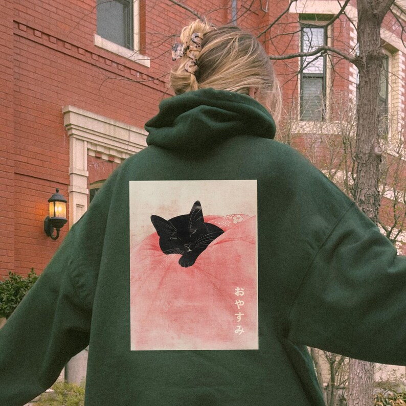 May include: A dark green hoodie with a black cat sleeping on a pink pillow. The cat is surrounded by flowers and the text "おやすみ" is printed on the pillow.