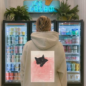 May include: A person wearing a beige hoodie with a black cat sleeping on a pink pillow printed on the back. The Japanese text "おやすみ" is printed below the cat. The person is standing in front of a refrigerator filled with ice cream.