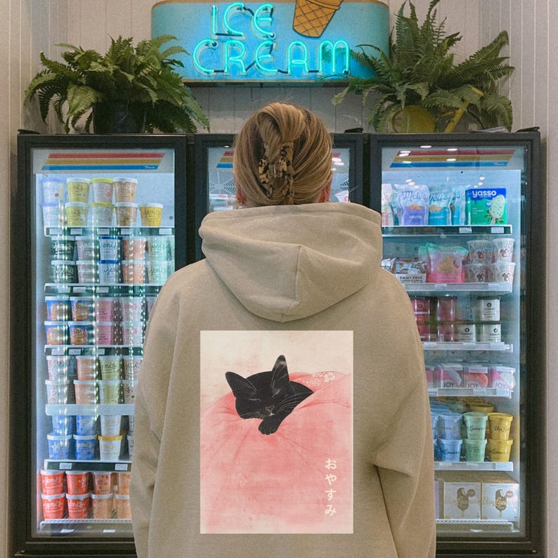 May include: A person wearing a beige hoodie with a black cat sleeping on a pink pillow printed on the back. The Japanese text "おやすみ" is printed below the cat. The person is standing in front of a refrigerator filled with ice cream.