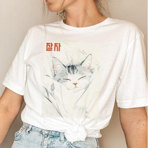 Peut inclure: T-shirt blanc avec un motif de chat dormant dans une couverture. Le texte "잠자" est imprimé en rouge sur le t-shirt.