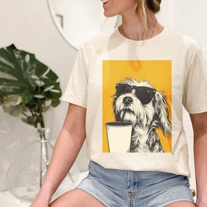 May include: A cream colored t-shirt with a graphic of a dog wearing sunglasses and holding a cup of coffee. The dog is on a yellow background.