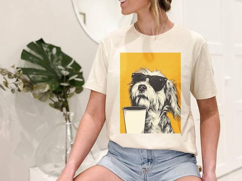 May include: A cream colored t-shirt with a graphic of a dog wearing sunglasses and holding a cup of coffee. The dog is on a yellow background.