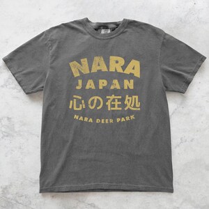 May include: Dark gray t-shirt with a distressed gold print. The print features the words "NARA JAPAN" in a bold font, along with Japanese characters and "NARA DEER PARK". The shirt has a classic crew neck and short sleeves.