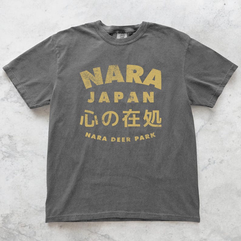 May include: Dark gray t-shirt with a distressed gold print. The print features the words "NARA JAPAN" in a bold font, along with Japanese characters and "NARA DEER PARK". The shirt has a classic crew neck and short sleeves.