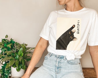 Puede incluir: Camiseta blanca con un gráfico de un gato negro y el texto japonés "こんちは", que se traduce como "hola".