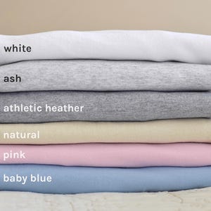 May include: A stack of six t-shirts in different colors, including white, ash, athletic heather, natural, pink, and baby blue.