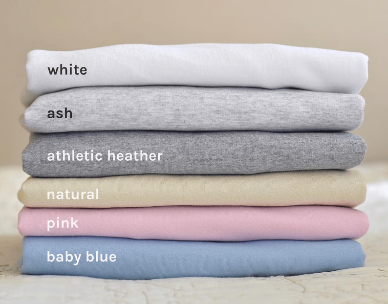 May include: A stack of six t-shirts in different colors, including white, ash, athletic heather, natural, pink, and baby blue.