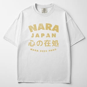 May include: White t-shirt with a distressed gold graphic. The graphic includes the word "NARA" above "JAPAN", Japanese characters, and "NARA DEER PARK". The shirt has a crew neck and short sleeves.
