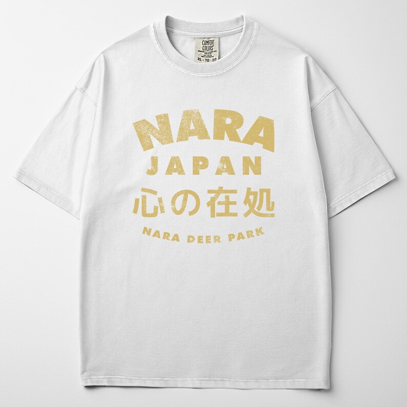 May include: White t-shirt with a distressed gold graphic. The graphic includes the word "NARA" above "JAPAN", Japanese characters, and "NARA DEER PARK". The shirt has a crew neck and short sleeves.