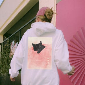 May include: White hoodie with a black cat sleeping on a pink pillow. The text "おやすみ" is printed on the hoodie.