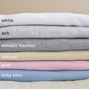 May include: Stack of six t-shirts in different colors, from top to bottom: white, ash, athletic heather, natural, pink, baby blue.