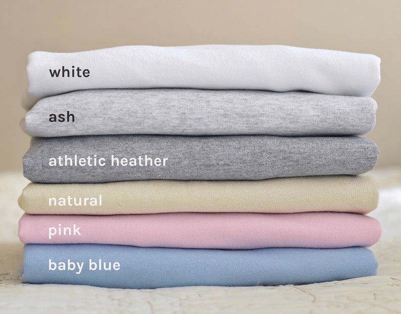 May include: Stack of six t-shirts in different colors, from top to bottom: white, ash, athletic heather, natural, pink, baby blue.
