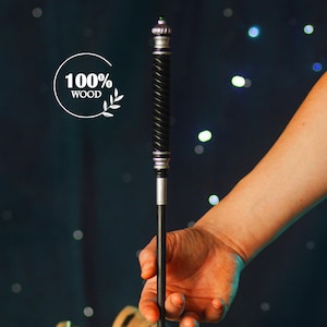 May include: A black and silver wand with a twisted design, held by a hand against a dark blue background with white lights. The wand is resting on top of rolled up parchment paper with a feather quill nearby. The text "100% WOOD" is visible in a white circle.