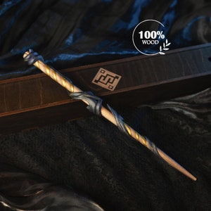 Op de afbeelding: Een houten toverstaf met een lichtbruine schacht, versierd met gouden en donkerblauwe details. De toverstaf wordt gepresenteerd in een donkere houten kist met een decoratief embleem. De tekst "100% WOOD" is zichtbaar.