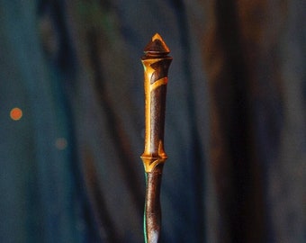 May include: A dark brown wooden wand with gold accents and a pointed tip. The wand is twisted and has a unique design. It is resting on a table with a feather and some scrolls.