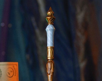 May include: A white and gold wand with a green gem on top. The wand is resting on a table with a book and a jar in the background.