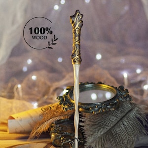May include: A wooden wand with a twisted, light brown handle and a gold-painted, intricate design. The wand is resting on a decorative mirror and a feather.