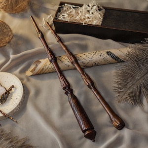Puede incluir: Dos varitas de madera con detalles intrincados, que descansan sobre una superficie de tela marrón con un trozo de pergamino enrollado y una pluma.