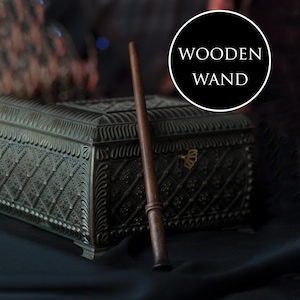 Puede incluir: Una varita de madera marrón oscuro con un diseño cónico, apoyada contra una caja de metal decorativa. La caja es oscura con patrones intrincados. El texto "WOODEN WAND" está en un círculo blanco.