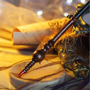 May include: A handcrafted wooden wand with a dark brown finish and a red tip, resting on a circular stone surface. The wand is detailed with carved patterns. A scroll and a decorative bottle are in the background.