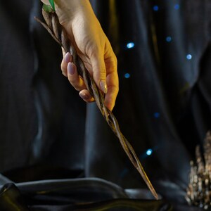 May include: A hand holds a dark brown, twisted wooden wand with a green leaf at the top. The wand is held against a black background.