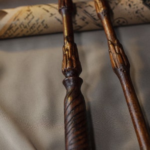 May include: Three wooden wands with intricate carvings and a dark brown finish. The wands are lying on a beige fabric surface.