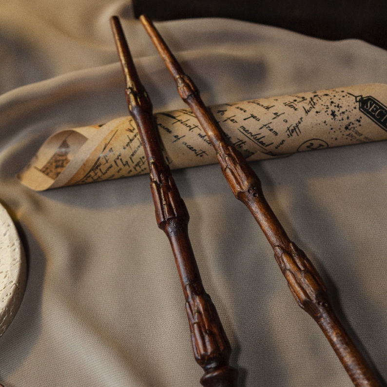 May include: Three wooden wands with intricate carvings, lying on a brown fabric surface with a rolled up piece of paper with handwritten text.