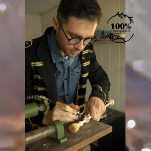 May include: A craftsman working on a wooden object with a rotary tool. The image shows a close-up of the artisan's hands and the object being carved. The text "100% HANDMADE" is visible in a circular logo.