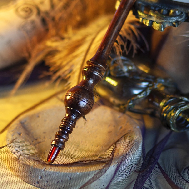 May include: A dark brown wooden pen with a red tip, featuring a detailed, carved design. The pen is set against a blurred background with feathers and a decorative element, creating a vintage aesthetic.