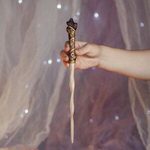 May include: A wooden wand with a twisted, light brown shaft and a dark brown, gold-accented handle with intricate carvings. The wand is held in a hand against a blurred background.