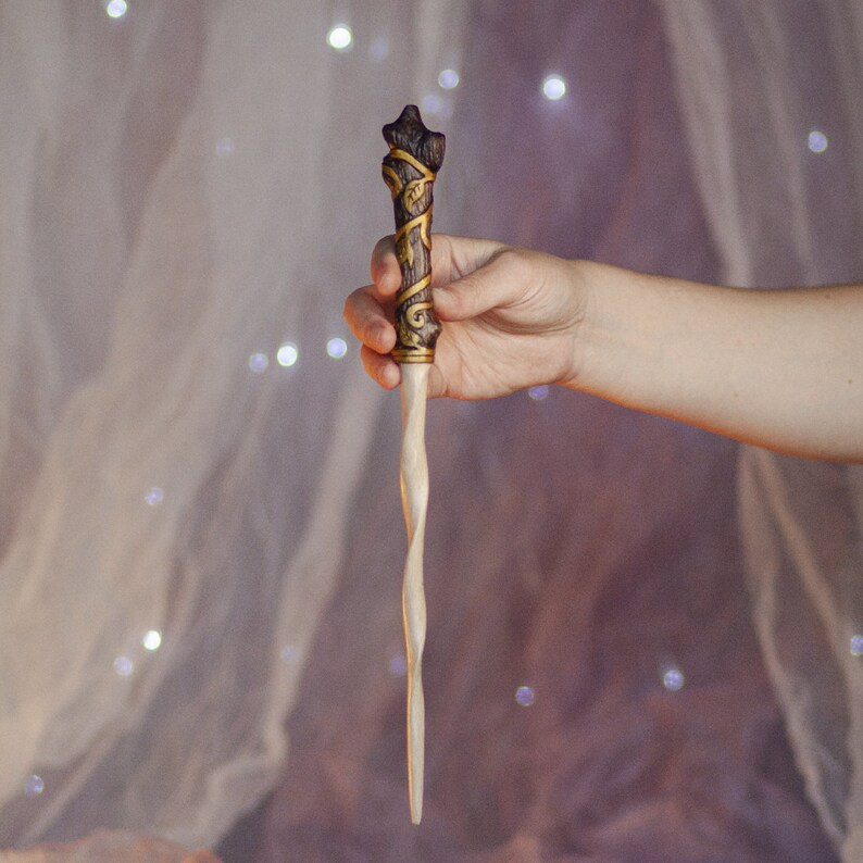 May include: A wooden wand with a twisted, light brown shaft and a dark brown, gold-accented handle with intricate carvings. The wand is held in a hand against a blurred background.