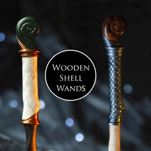 May include: Two wooden wands with spiral shell-shaped tops. The left wand has a green top, a white handle with a cross-hatched pattern, and a gold ring. The right wand has a brown top and a dark grey handle with a cross-hatched pattern. Text reads: WOODEN SHELL WANDS.
