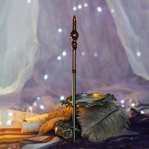 May include: A detailed wooden wand with a dark brown handle and a black shaft, topped with a red tip. The wand is displayed with a decorative golden dish and feathers, set against a soft, blurred background with twinkling lights.