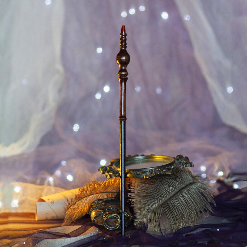 May include: A detailed wooden wand with a dark brown handle and a black shaft, topped with a red tip. The wand is displayed with a decorative golden dish and feathers, set against a soft, blurred background with twinkling lights.