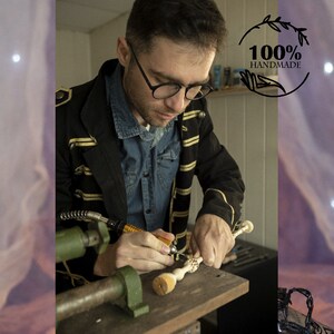May include: A person is carving a wooden wand with a small electric tool. The wand has intricate details and is made of light-colored wood. The person is wearing a black jacket with gold trim and a blue denim shirt. The background is a workshop with tools and other items.