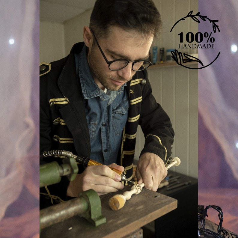 May include: A person is carving a wooden wand with a small electric tool. The wand has intricate details and is made of light-colored wood. The person is wearing a black jacket with gold trim and a blue denim shirt. The background is a workshop with tools and other items.