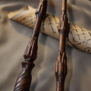 May include: Three wooden wands with intricate carvings and a gold-colored finish. The wands are lying on a rolled-up piece of parchment paper.