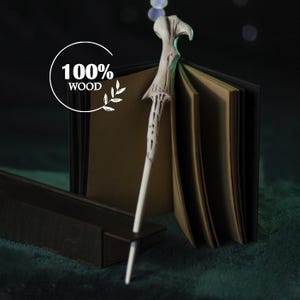 Puede incluir: Una varita mágica de madera hecha a mano con un mango detallado en forma de asta, apoyada contra un libro abierto. La varita es blanca con detalles marrones. La imagen incluye el texto "100% WOOD" en un círculo blanco.